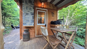 A wooden cabin with a stone wall, a bench, a table with glasses and a bottle, and a door with a window. - Waterfront Glamping | La Nomade (Shannon)