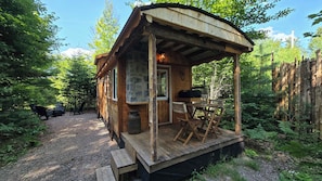 A rustic cabin with a porch, a table, and chairs, surrounded by dense forest. - Waterfront Glamping | La Nomade (Shannon)