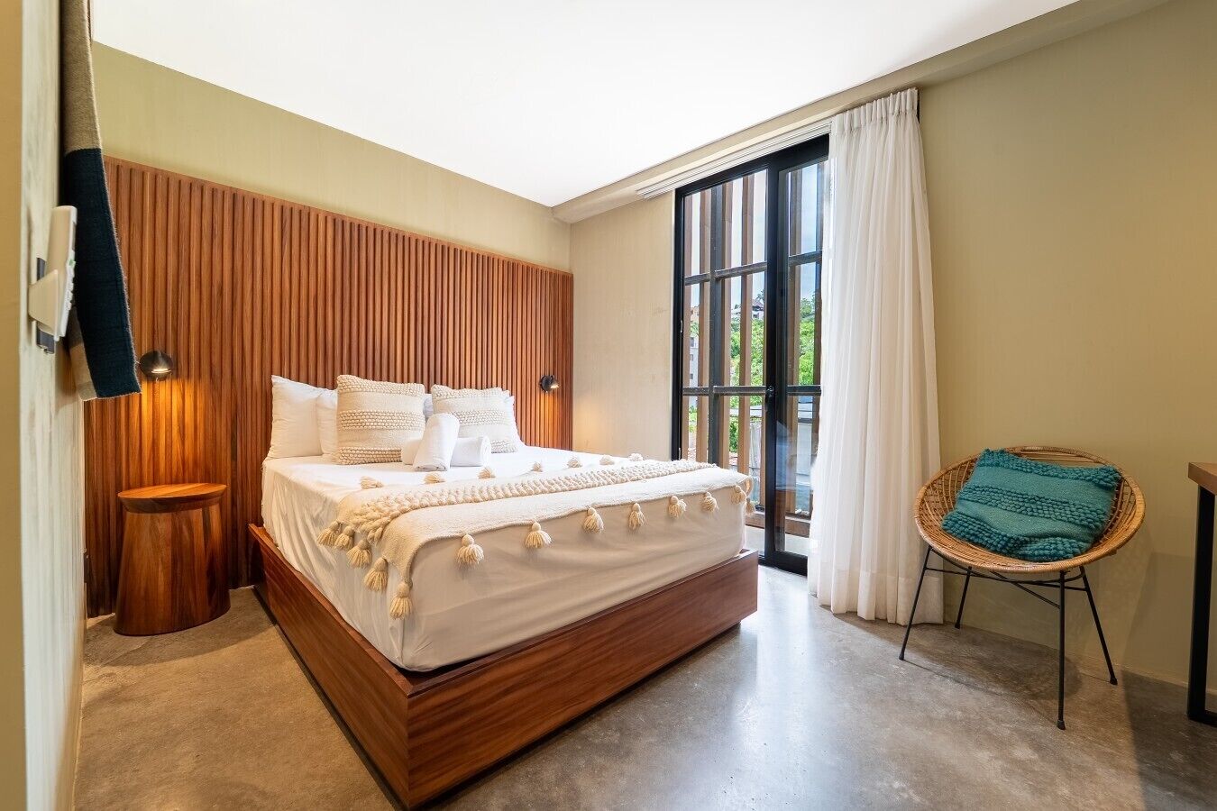 A bedroom with a large bed, a wooden headboard, a small side table, a wicker chair, and a view through a glass door.