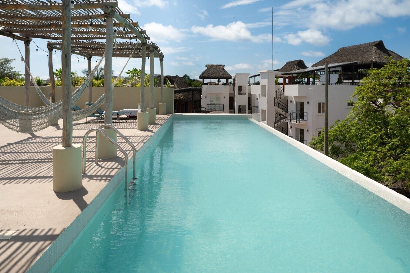 Una piscina con una pérgola y hamacas, rodeada de edificios con techos de paja.