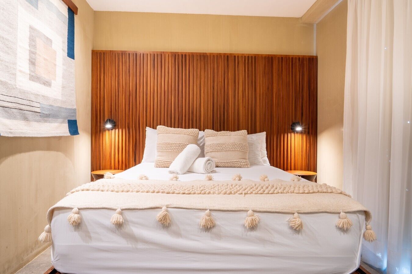 Ein Bett mit Holz-Kopfteil, zwei Nachttischlampen und einer weißen Bettdecke mit Fransen.