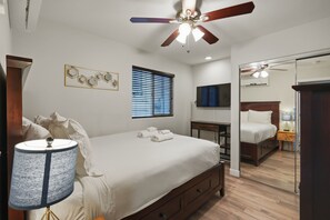 A bedroom with a bed, a ceiling fan, a window with blinds, a wall-mounted TV, and a mirror.