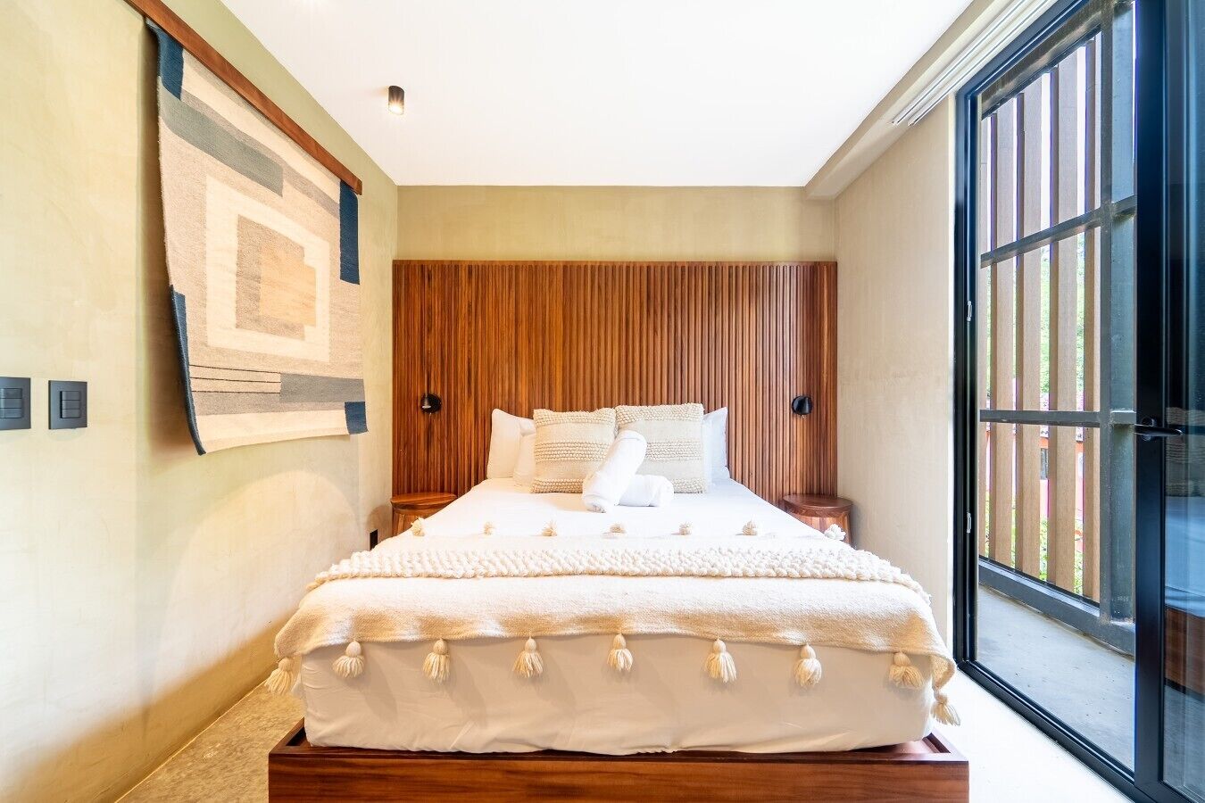 Une chambre à coucher avec une tête de lit en bois, un grand lit avec une literie blanche et une fenêtre équipée de stores verticaux.