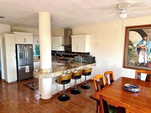 A kitchen with a central island, bar stools, a dining table, and a painting on the wall. - Private House & Casita with Pool and Ocean Views (Cruz de Huanacaxtle)