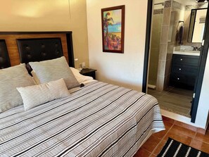 A hotel room with a bed, a nightstand, a painting, and a bathroom visible through an open door. - Private House & Casita with Pool and Ocean Views (Cruz de Huanacaxtle)