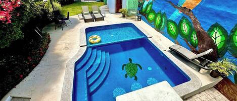 Une piscine ornée d’une fresque représentant un perroquet et une tortue, entourée de chaises longues et d’un petit bâtiment.