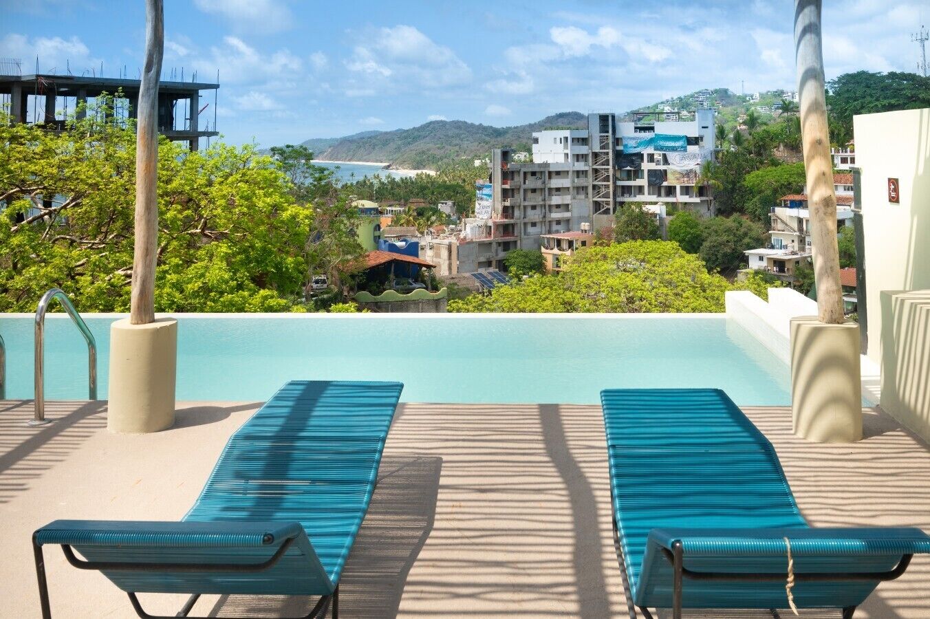 A pool area with two lounge chairs, a clear view of the sea, and surrounding buildings.