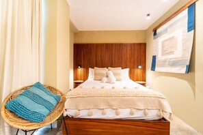 A hotel room with a bed, a nightstand, a wall-mounted lamp, a wicker chair, and a wall art piece. - Modern Studio | Rooftop Pool + Parking (Sayulita)