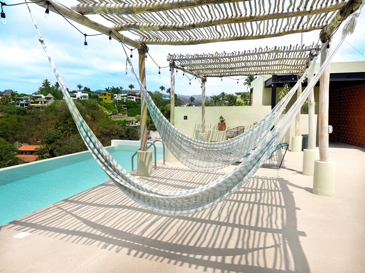 A hammock under a pergola with a pool and a view of houses in the background.