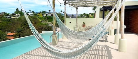 Uma rede sob uma pérgola, com piscina e vista para casas ao fundo.