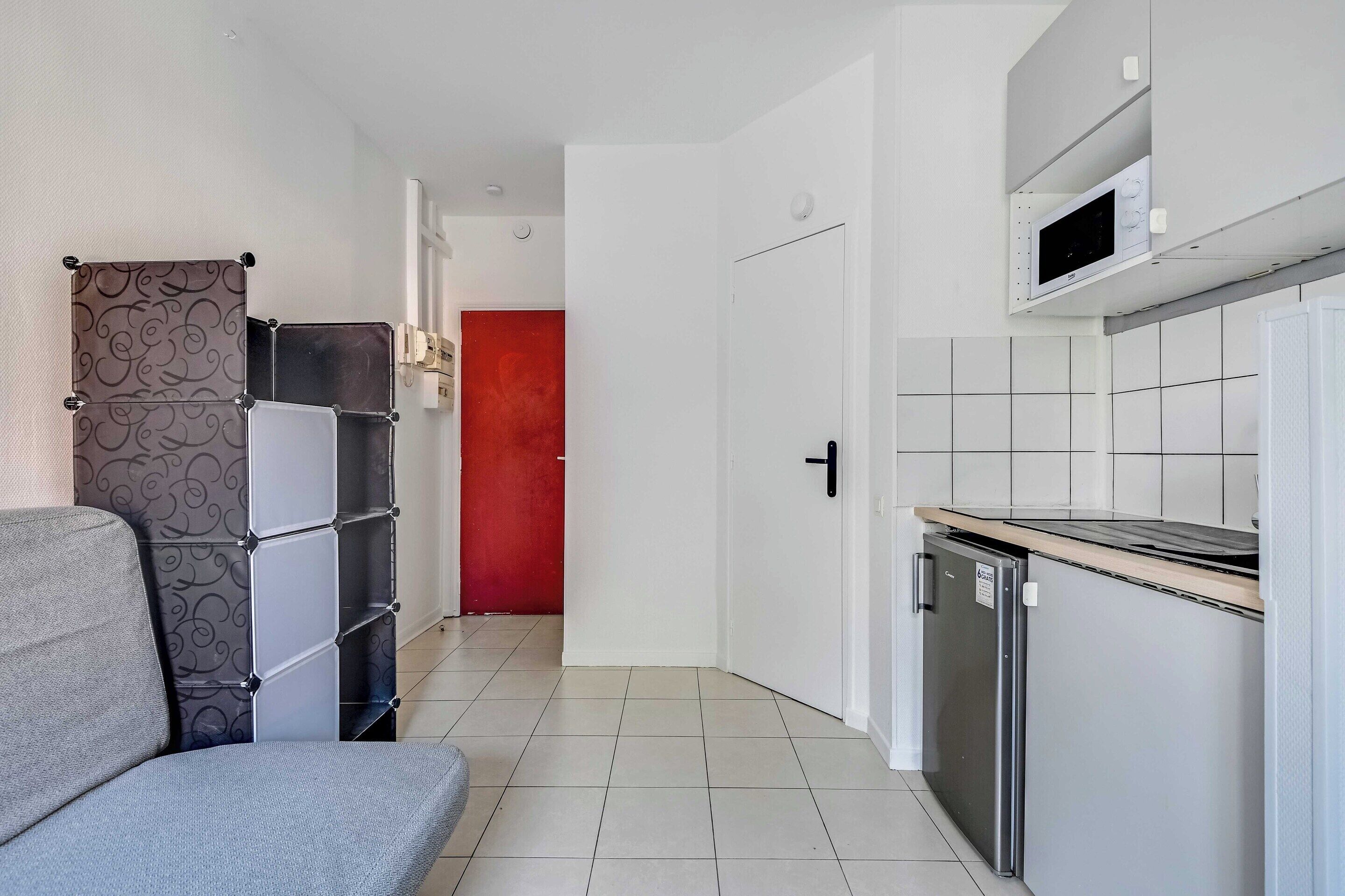 A compact kitchen with white cabinets, a grey sofa, and a red door in the background.