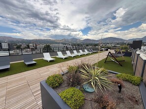 A rooftop lounge area with sun loungers, a wooden deck, and a view of a cityscape and mountains. - Prime Location Rooftop, Pool, Hot Tub & Gym (Salt Lake City)