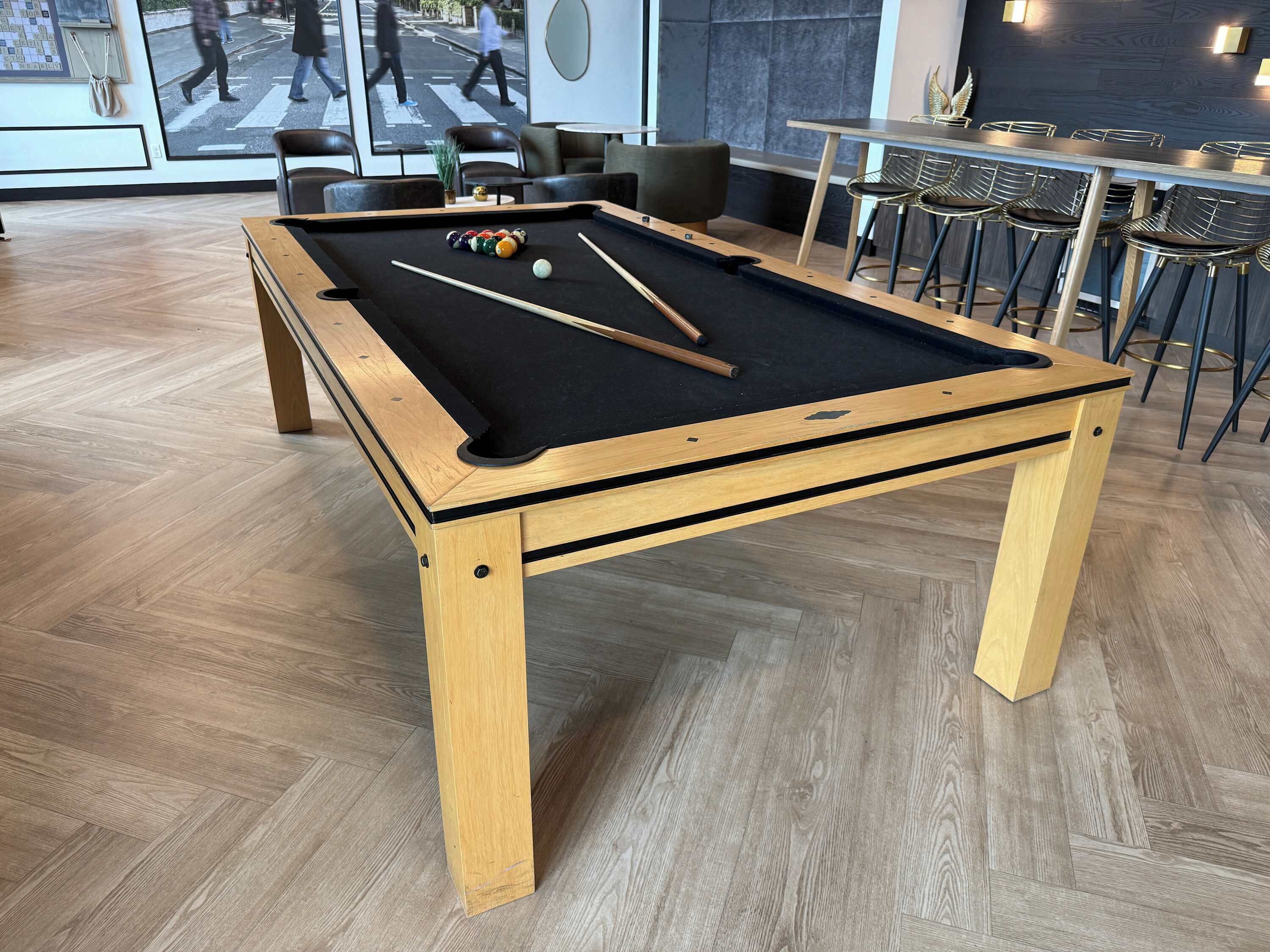 A pool table with balls and cues, surrounded by chairs and tables in a room with wooden flooring.