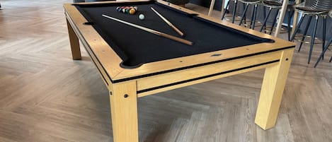 A pool table with balls and cues, surrounded by chairs and tables in a room with wooden flooring.