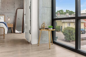 A modern room with a large window, a bed, a bedside table with a lamp, and a small table with a plant. - Prime Location Rooftop, Pool, Hot Tub & Gym (Salt Lake City)