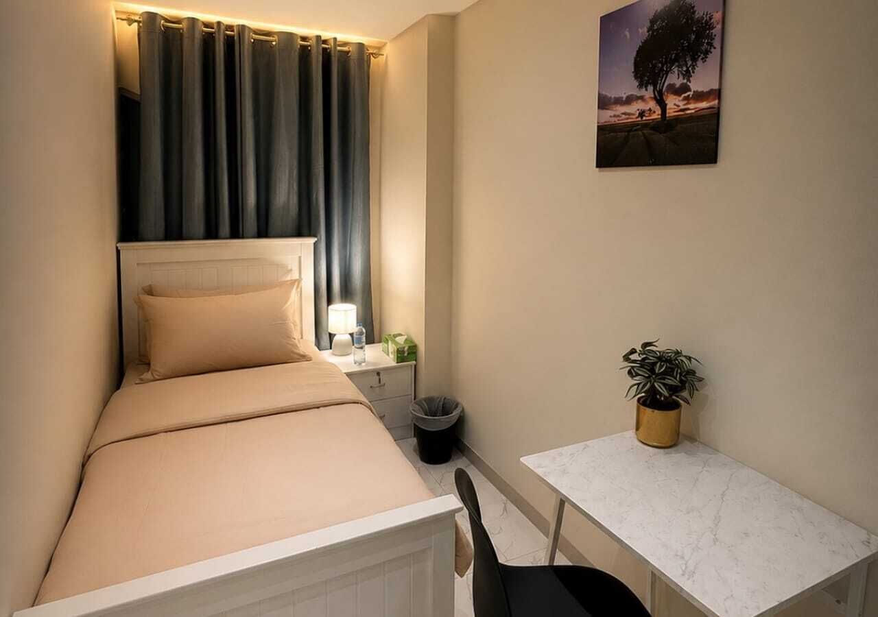 Een compacte kamer met een bed, nachtkastje, lamp, prullenbak en een kleine tafel met een plantje.