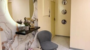 A modern interior with a marble wall, a round mirror, a small round table, two grey chairs, and a shelf with decorative items.