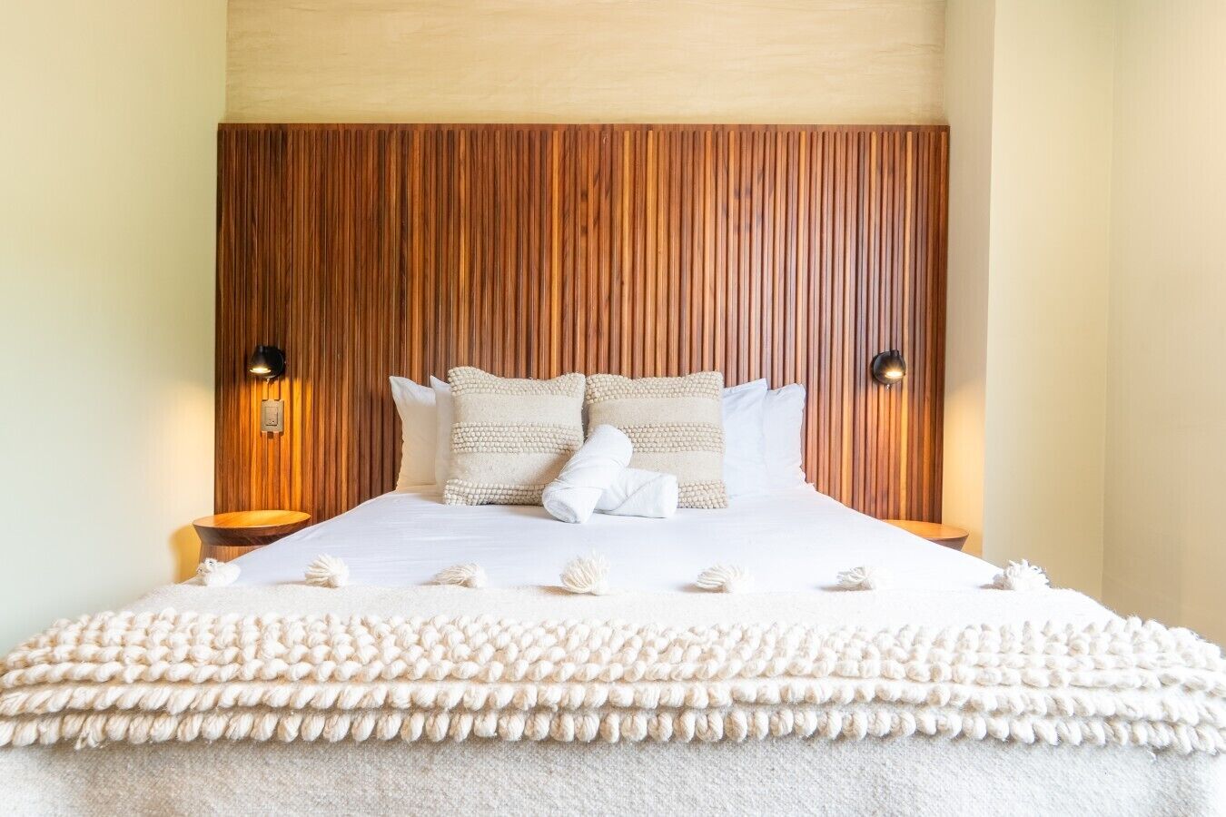 Un lit bien fait, recouvert d’une housse blanche à texture intéressante, avec deux lampes de chevet et une tête de lit en bois.