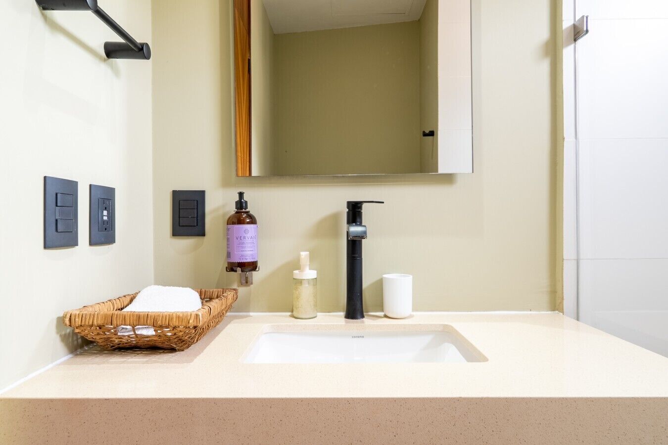 A bathroom with a white sink, a black faucet, a bottle of liquid soap, a white cup, and a basket of white towels.