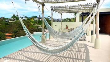 Una hamaca bajo una pérgola con piscina y vistas a casas al fondo.
