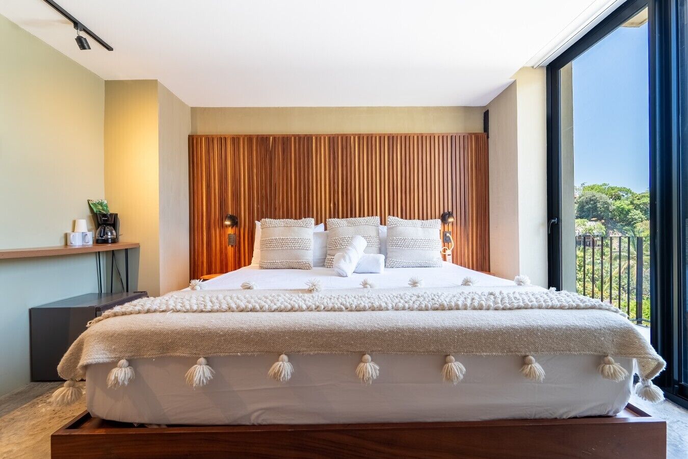 A modern bedroom with a large bed, wooden headboard, and a view of greenery through a balcony.