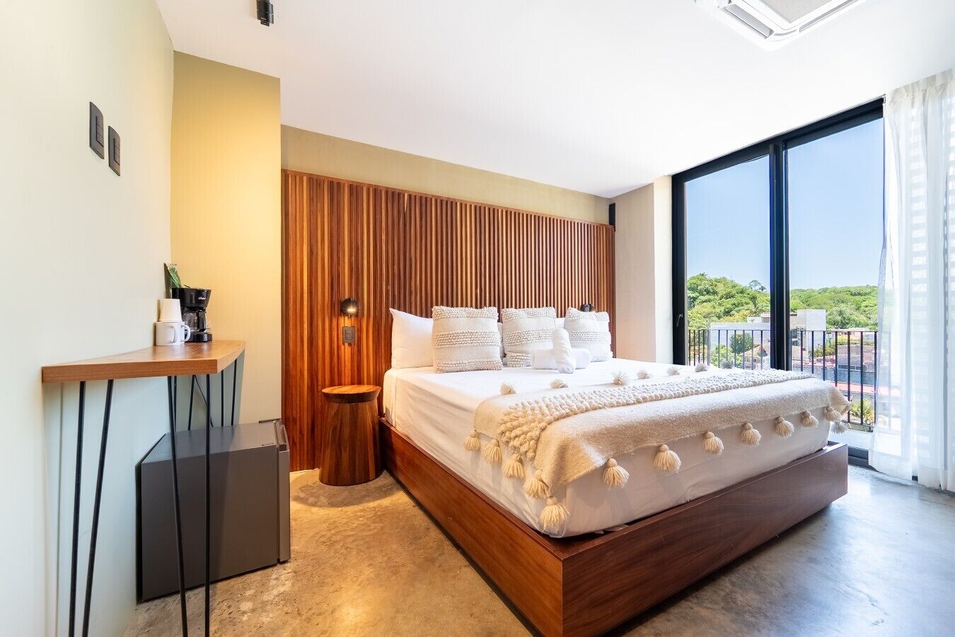 Un dormitorio moderno con una cama grande, cabecera de madera, una mesita de noche con cafetera y vistas al exterior a través de un ventanal amplio.