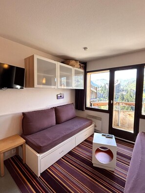 Een knus ingerichte kamer met een bank, een klein tafeltje, een aan de muur bevestigde televisie en een raam met uitzicht op gebouwen en bergen.