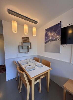 Ein Essbereich mit einem Tisch für vier Personen, Holzstühlen, Pendelleuchten, einem an der Wand befestigten Flachbildfernseher und einem gerahmten Bild über dem Tisch.