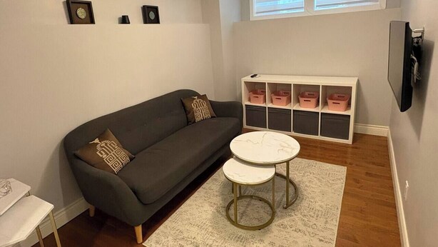 A living room with a sofa, two small tables, a white rug, a white storage unit with pink bins, and a TV mounted on the wall.