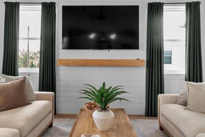 Ein Wohnzimmer mit einem Wand-Fernseher, zwei Sofas, einem Couchtisch mit Pflanze und grünen Vorhängen.