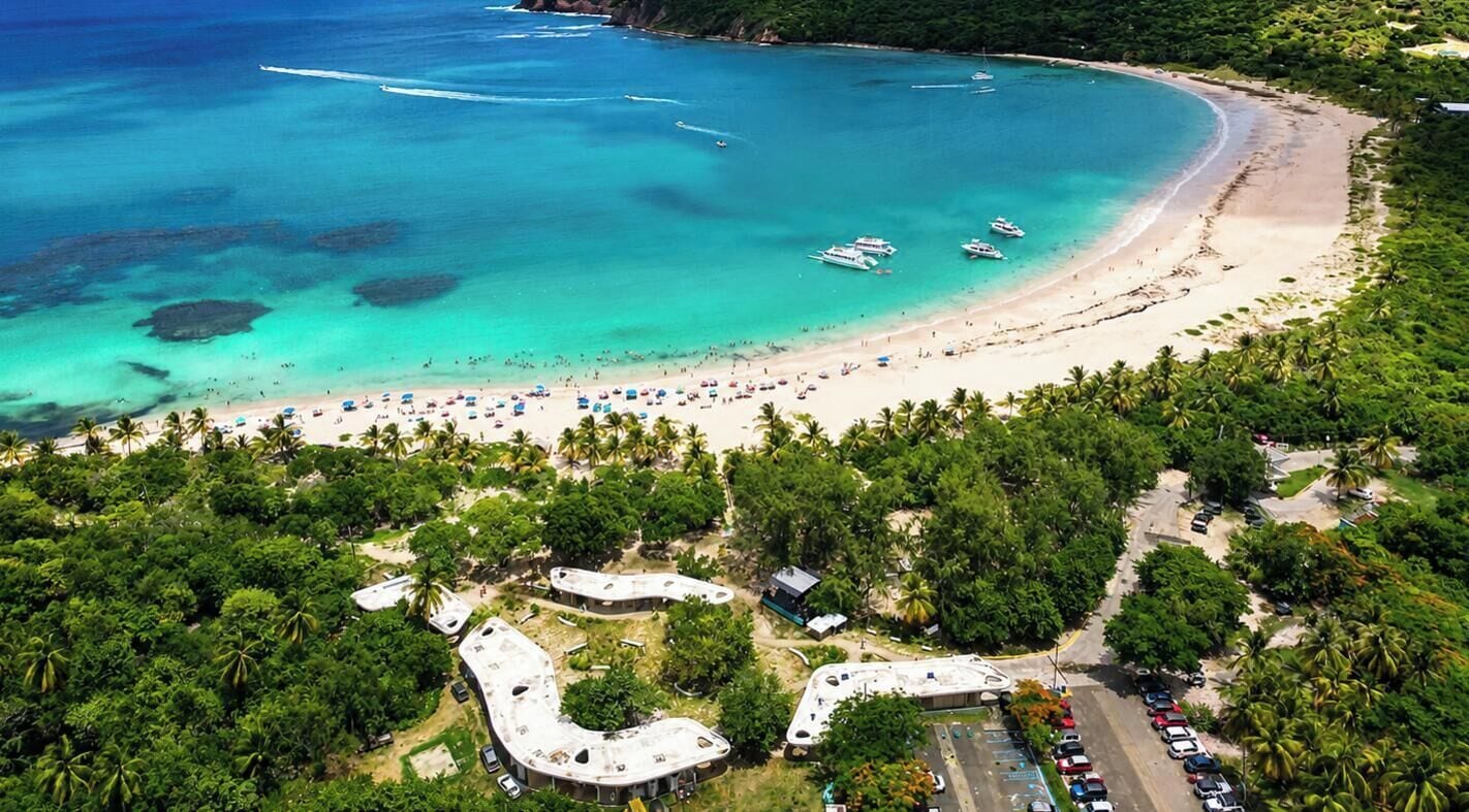An aerial view of a beach with boats, a sandy shore, and a parking area with cars.