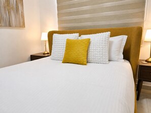 Ein ordentlich bezogenes Bett mit weißer Bettwäsche und zwei Kissen, eines gelb und eines weiß, in einem Zimmer mit gestreifter Wand und einer Lampe auf einem Nachttisch.