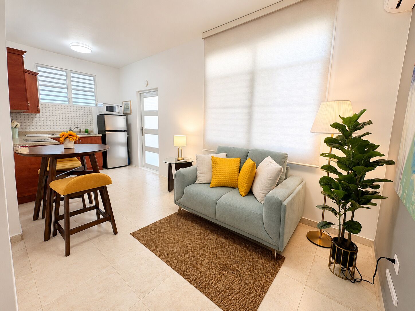 A living room with a sofa, a dining area with chairs, a kitchen with appliances, and a potted plant.