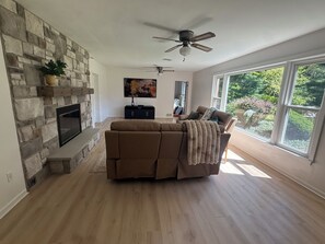 A living room with a stone fireplace, a ceiling fan, and large windows.