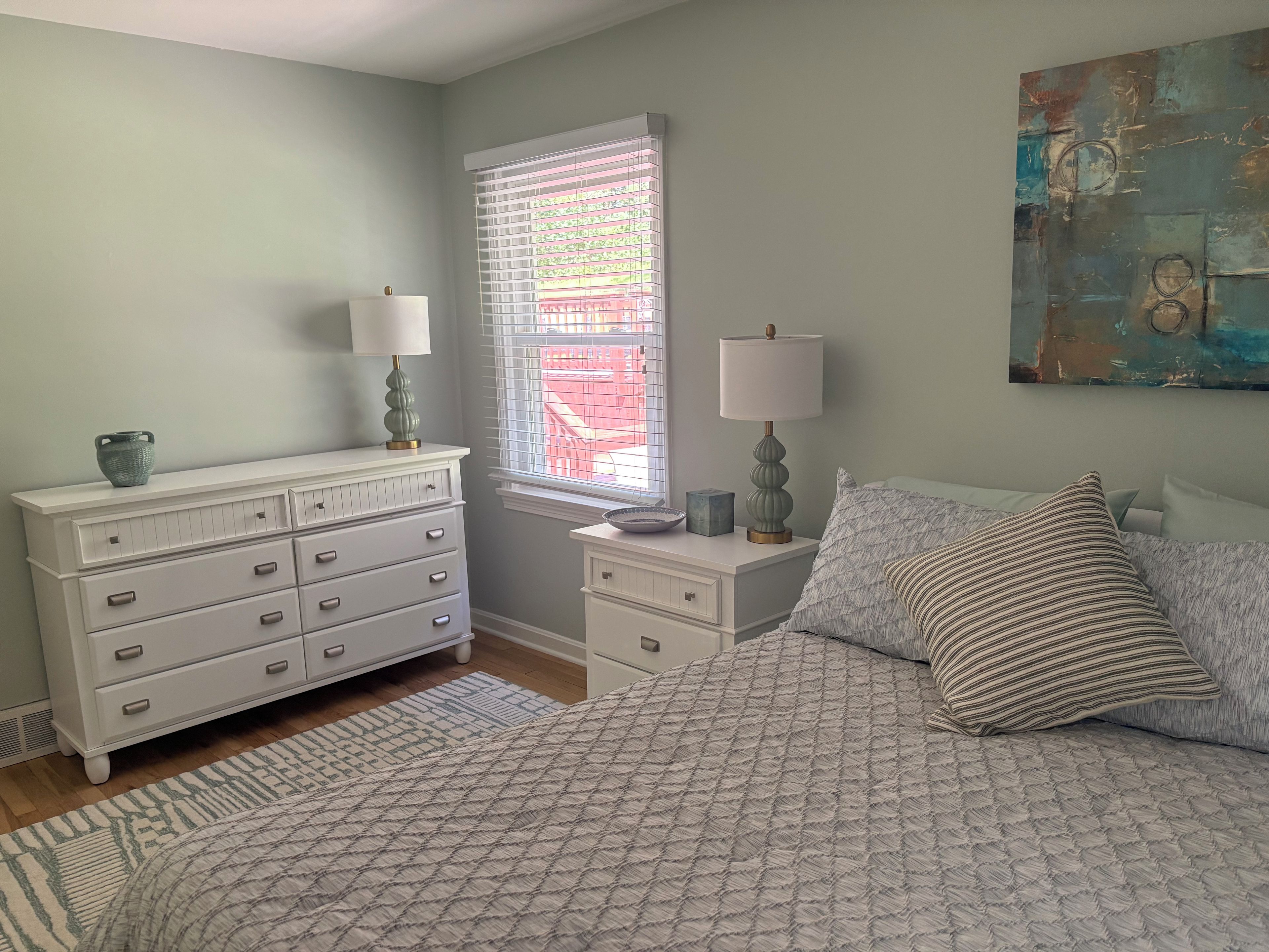 Une chambre à coucher comprenant un lit, une commode, deux tables de chevet, une fenêtre avec des stores et un tableau accroché au mur.