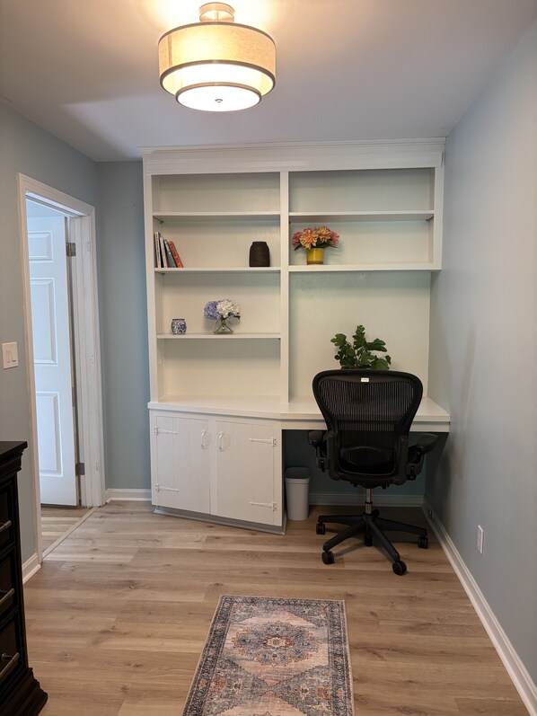 A room with a white built-in desk, a black office chair, and a patterned rug on a wooden floor.
