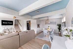 A spacious living area with a sofa, a dining table with chairs, a kitchen in the background, and a staircase leading to an upper level. - Cape May Escape • Sleeps 10 • Tiki Bar • Grill (Cape May)