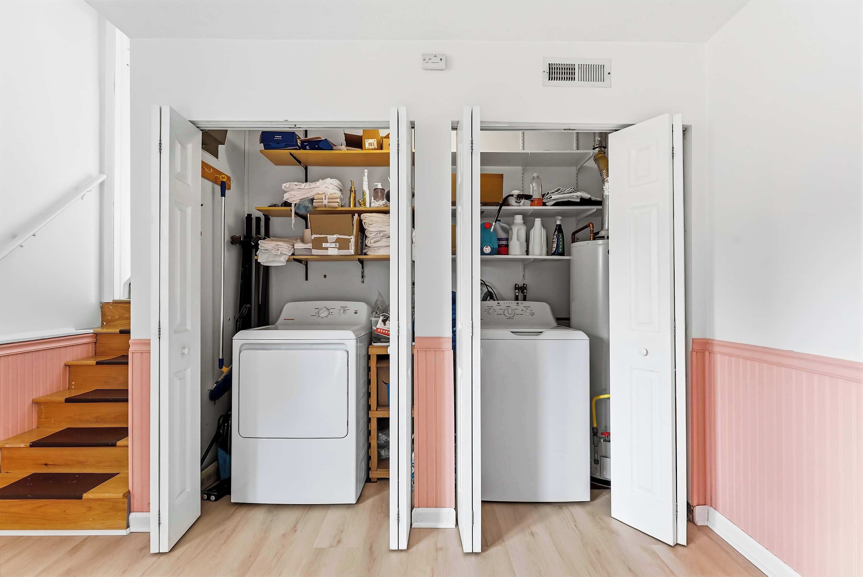 A laundry room with two washing machines, shelves with cleaning supplies, and a wooden staircase.