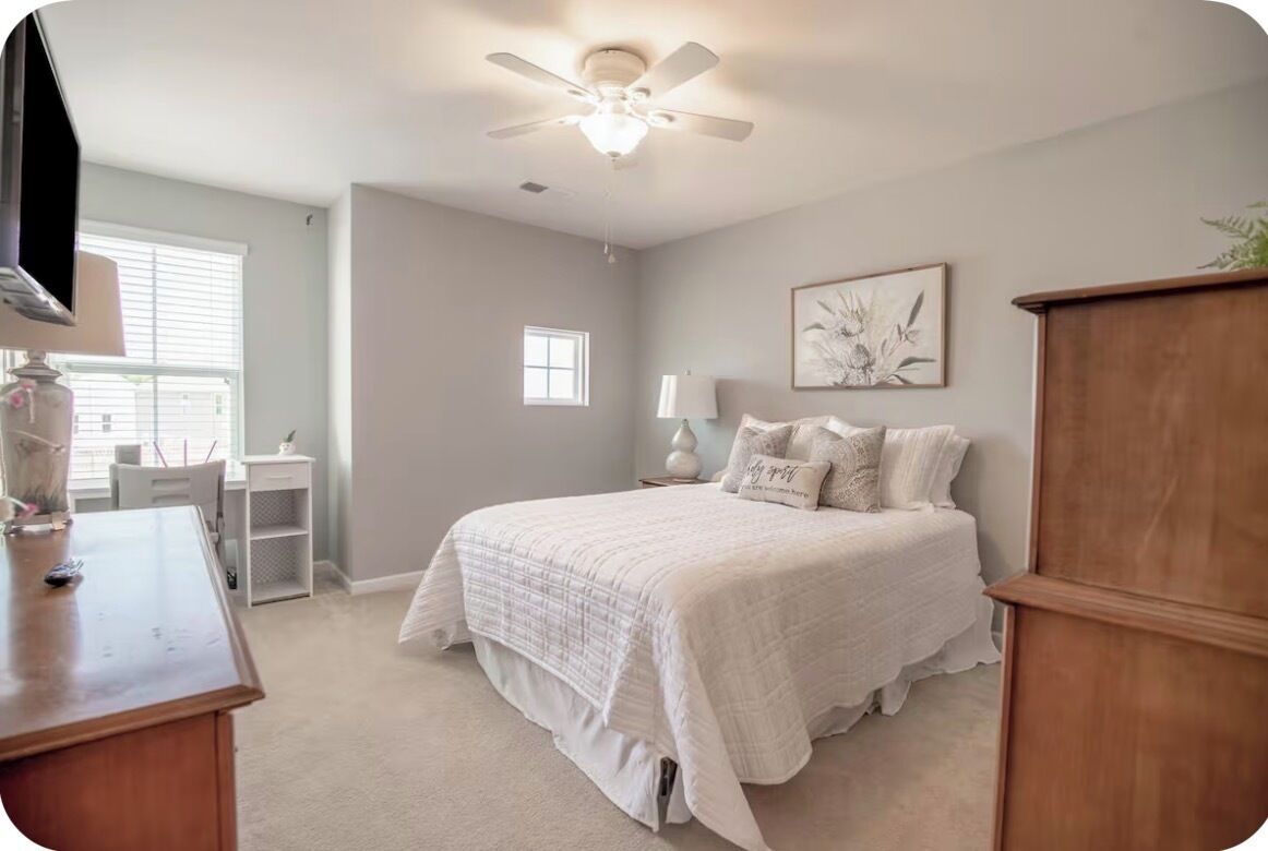 A bedroom with a bed, a desk, a chair, a ceiling fan, and a window.