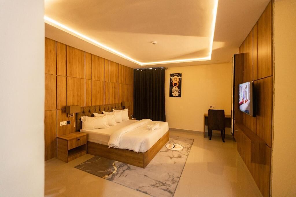 A modern bedroom with a wooden headboard, a flat-screen TV mounted on the wall, a bedside table, and a small desk with a chair.