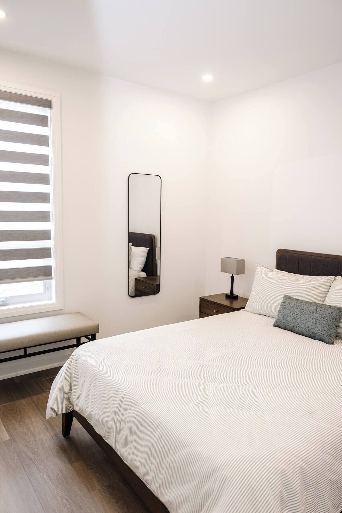 A neatly arranged bedroom with a bed, bedside table, lamp, and a mirror on the wall.