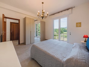 Quarto com cama grande, lustre, varanda com vista e porta para outro cômodo.