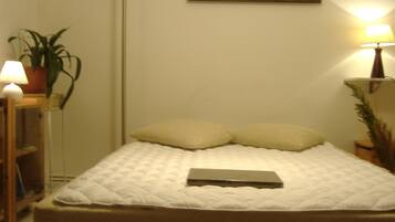A neatly made bed with a quilted cover and two pillows, a bedside lamp, a small table with a plant, and a painting on the wall.