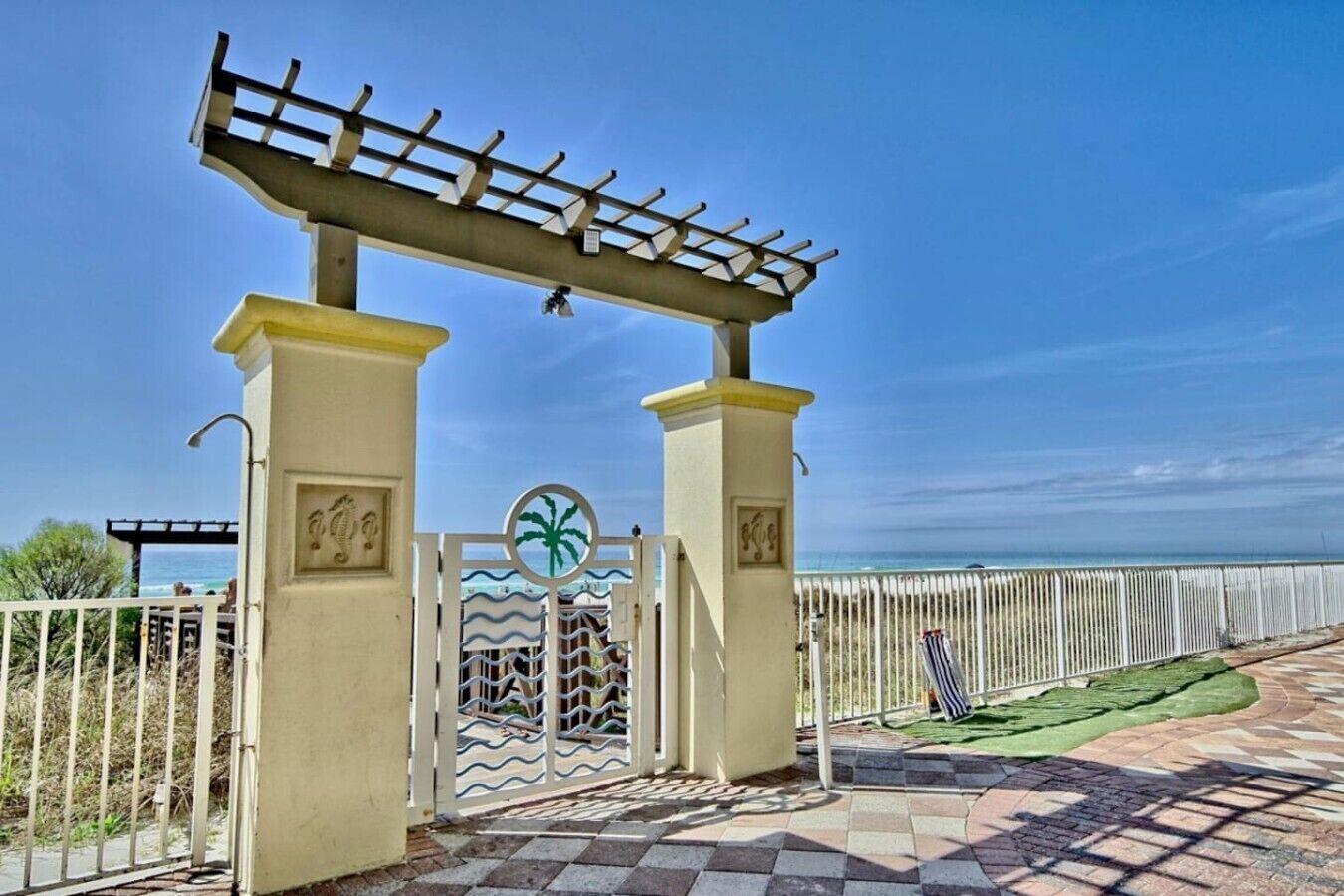 Een toegangshek met een pergola erboven, een palmboom symbool op het hek, en uitzicht op zee.