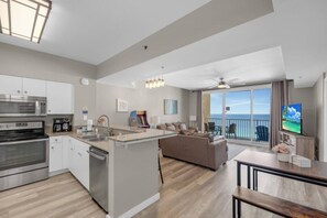 A modern kitchen with a central island, a dining area with a TV, and a balcony with a view of the ocean. - 2-Bedroom Beachfront Condo at Shores of Panama! (Panama City Beach)