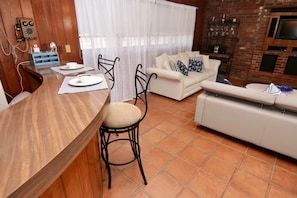 A dining area with a wooden table and chairs, a white sofa, and a TV mounted on a brick wall. - Historic Pool House on 4-acre Mango Farm in heart of Miami-Reduced this Summer (Miami)