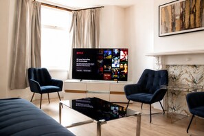 A modern living room with a flat-screen TV displaying a Netflix interface, two dark blue armchairs, a glass coffee table, and a marble fireplace. - Contractors or Families | Sleeps 12 + Fast WiFi | Near Bradford University + BRI (Bradford)