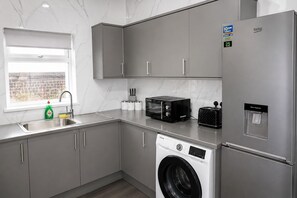 A modern kitchen with grey cabinets, a stainless steel refrigerator, a built-in washing machine, a toaster, and a water dispenser. - Contractors or Families | Sleeps 12 + Fast WiFi | Near Bradford University + BRI (Bradford)