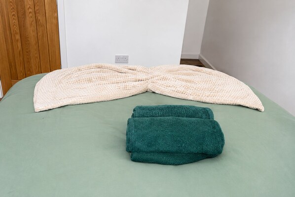 Een bed met een groen laken en twee opgevouwen dekens, een beige en een blauwgroene.