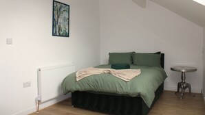 A bedroom with a bed, a radiator, a small table, and a painting on the wall. - Stylish Bradford Home | Near City Centre | Sleeps 11 (Bradford)